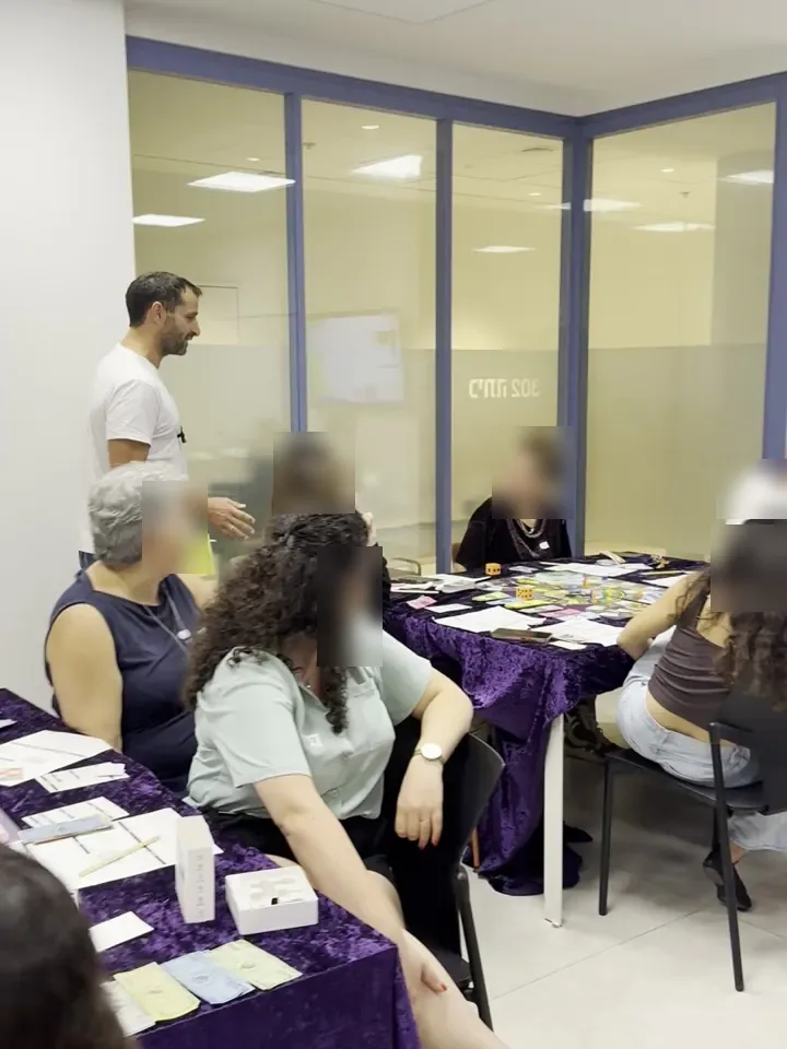 משתתפים יושבים סביב שולחן משחק, ואלון בר עומד לידם ומנחה